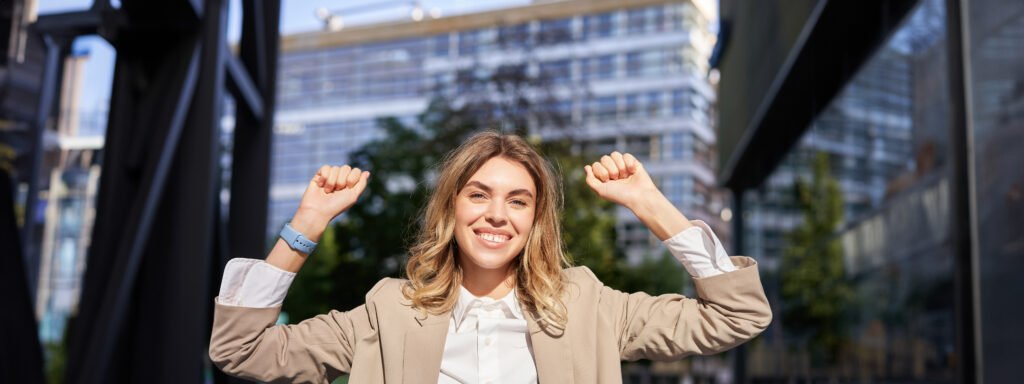 Happy smiling saleswoman in beige suit, celebrates her achievement, triumphing, making fist pump and looking excited on street of business center