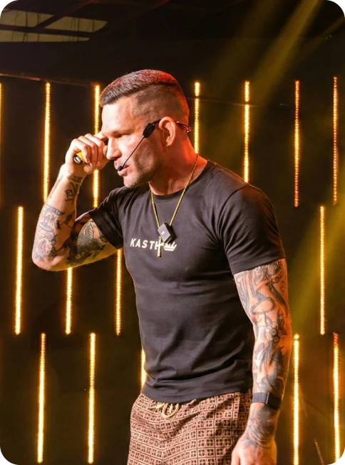 Adri Kastel impartiendo una formación de alto impacto sobre un escenario con iluminación técnica. El emprendedor viste ropa de marca propia y porta un micrófono de diadema, proyectando una imagen de autoridad, éxito y mentoría premium. Fondo con barras de luz cálida que refuerzan el branding corporativo en tonos dorados y negros, ideal para representar la transformación y el estatus de sus programas de formación.